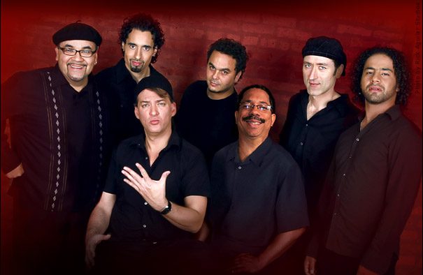 Seven men posing against a red background.